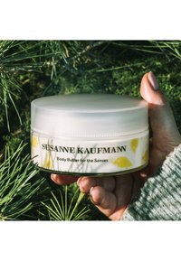 Un pot blanc de beurre corporel Susanne Kaufmann avec des motifs d'oiseaux jaunes, un couvercle transparent et une étiquette imprimée. Une main tient le pot parmi le feuillage vert.