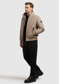 Beige bomberjakke med høy krage, to svarte lommer med glidelås, ribbestrikkede mansjetter og kant, kombinert med svarte bukser og svarte støvler.