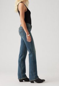 Femme aux longs cheveux blonds portant un haut noir sans manches, un jean bleu droit et des bottes noires à talons, debout de profil.