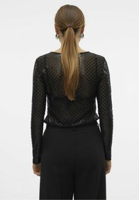 Kvinna med långt blont hår i hästsvans, iförd en svart genomskinlig långärmad topp med rutmönster och svarta högmidjade byxor, vänd bort.