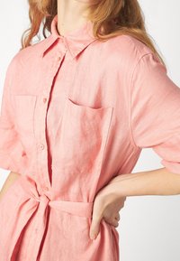 Camicia di lino rosa con colletto alla francese, maniche corte, due tasche sul petto e una cintura in vita. Texture liscia e leggera.