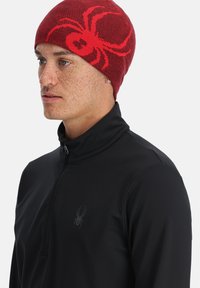 Berretto lavorato a maglia rosso con un design di ragno nero, abbinato a una maglietta nera con collo a zip. Il tessuto appare liscio e aderente.