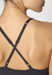 Des bretelles de soutien-gorge noires forment un motif en X dans le dos, avec un matériel coulissant réglable. Une peau lisse est visible sous les bretelles.