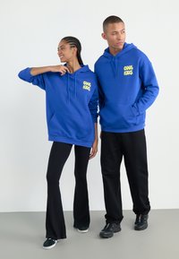 Blue hoodies with "COOL KIDS" print in yellow, featuring a front pocket, paired with black flared pants and sneakers, on a neutral background.