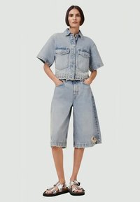 Camisa cropped de denim azul claro com mangas curtas, duas bolsos frontais, combinada com calções até ao joelho com um detalhe desgastado. Sandálias prateadas.
