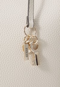 Sangle de sac en cuir texturé blanc avec des accents de clés et de charms en ton doré, comportant une étiquette rectangulaire embossée du mot "GUESS."
