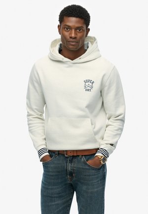 Cream hooded sweatshirt with a front pocket and navy logo. Features striped cuffs. Worn with blue denim jeans and a brown belt.