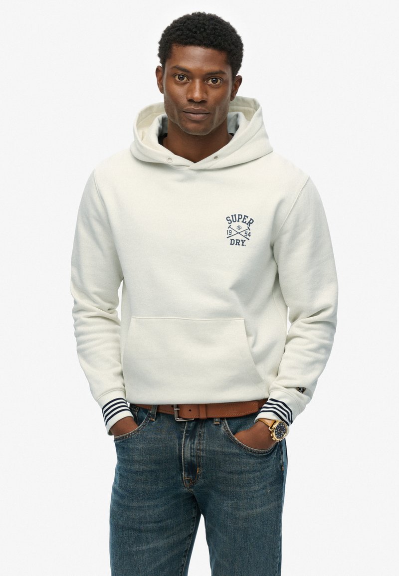 Kremowa bluza z kapturem z przednią kieszenią i granatowym logo. Ma paskowane mankiety. Nosi się z niebieskimi dżinsami i brązowym paskiem.