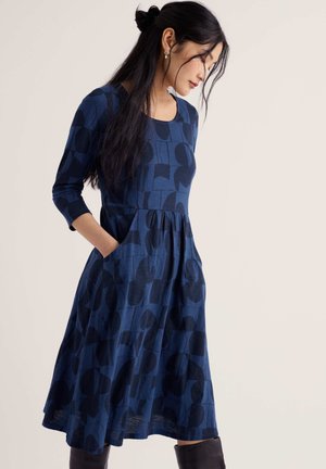 Woman with long dark hair wears blue patterned dress with three-quarter sleeves and black boots, standing against light background.