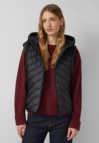 Junge Frau mit langen blonden Haaren, die eine schwarze gesteppte Kapuzenweste über einem burgunderfarbenen Pullover und dunklen Jeans trägt, steht vor einem schlichten Hintergrund.