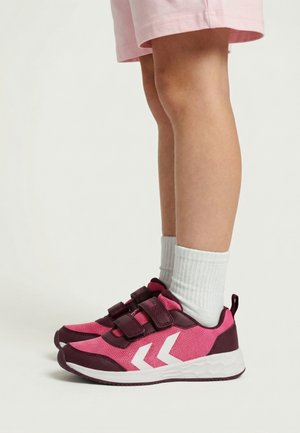 Bambino che indossa scarpe da ginnastica rosa e granata con strisce bianche a chevron, calzini bianchi e pantaloncini rosa chiaro, in piedi su uno sfondo neutro.