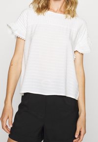 Bílá pruhovaná blůza s krátkými volánkovými rukávy a krajkovým zdobením. Sestaveno s černými šortkami, zdůrazňující uvolněný a neformální design.