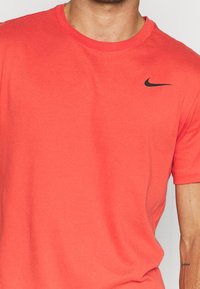 Mężczyzna w koralowo-czerwonej koszulce Nike z czarnym logo swoosh na prawym klatce piersiowej, z krótkimi rękawami i widocznym tatuażem na prawym przedramieniu.