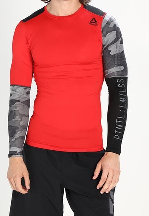 Homme portant un haut de compression rouge Reebok avec des manches longues à motif camouflage et noires, assorti à un short de sport noir.
