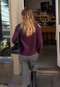 Overgrote paarse trui, grijze broek met fijne streep en wijde pijpen, en kniehoge zwarte laarzen. Zwarte handtas hangt over de schouder. Buiten cafésetting.