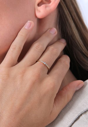 Goldring mit einem zentralen ovalen Stein, flankiert von kleineren runden Edelsteinen, auf einer Hand mit natürlicher Hauttextur gesetzt.