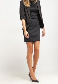 Czarna dopasowana sukienka z krótkim rękawem, w połączeniu z czarnym żakietem. Całość dopełniają szpilki. Gładka faktura na całej powierzchni.