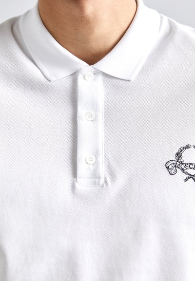Valge polo särk tekstureeritud kangast, kolme nupuga kaelusega ja musta värvi tikandiga logo kujundusega vasakul küljel.