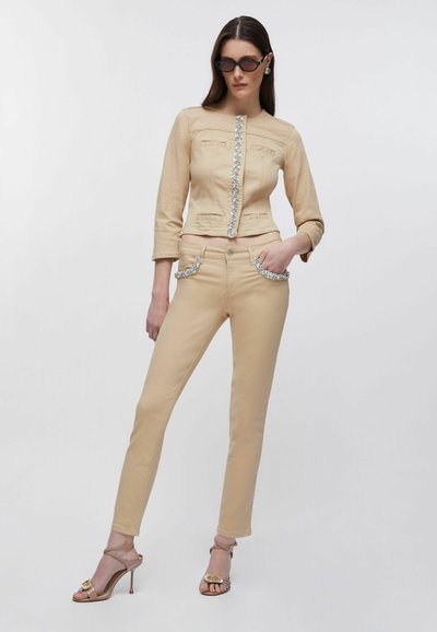 Femme portant un pantalon beige slim et une veste assortie ornée de strass, des sandales à talons hauts marron et des lunettes de soleil foncées, debout devant un fond blanc.