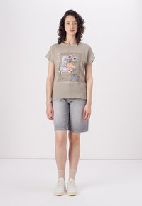 T-shirt gris clair avec impression graphique florale, manches courtes et col rond, associé à un short en jean de longueur genou et des baskets légères.