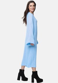 Abito maglione azzurro chiaro a maniche lunghe, con polsini a coste e spacchi laterali, indossato con stivali neri con tacco a piattaforma.