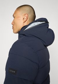 Mørkeblå dunjakke med hette, med en glatt tekstur, quiltet design og svarte detaljer. Fôret med lys grå materiale.
