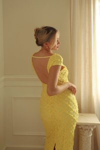 Robe en dentelle jaune avec texture florale, silhouette ajustée, dos ouvert, manches courtes et fente latérale, mise en valeur par un éclairage doux et élégant.