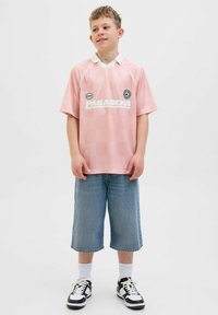 Camiseta de fútbol de manga corta color rosa claro con cuello blanco y logo, combinada con pantalones cortos de mezclilla azules y holgados, y zapatillas blancas y negras.