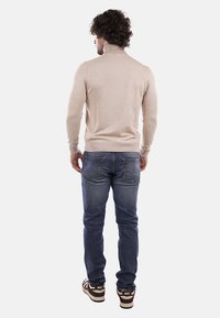Maglione beige a collo alto, design aderente, polsini e orlo a coste, abbinato a jeans blu. Il soggetto è in piedi con la schiena rivolta verso l'osservatore.
