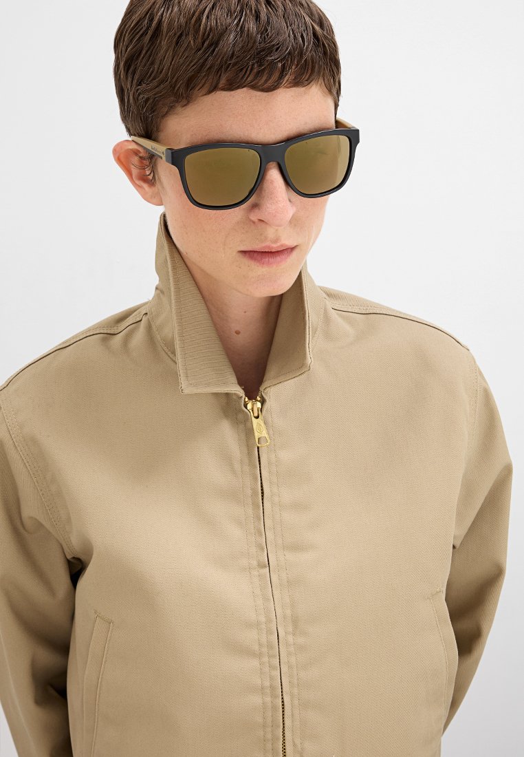 Beżowa kurtka zapinana na zamek z wysokim kołnierzem, wykonana z wytrzymałego materiału. Czarne okulary przeciwsłoneczne z brązowymi szkłami tworzą nowoczesny wygląd.