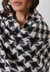 Geruit sjaal in zwart en crème, gemaakt van zacht materiaal. Heeft een afwisselend patroon van grote en kleine vierkanten en valt losjes.