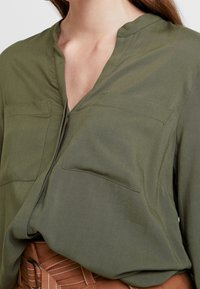 Blusa verde oliva con scollatura a V, dotata di due tasche frontali, realizzata in materiale leggero e con una vestibilità rilassata, abbinata a una cintura marrone.