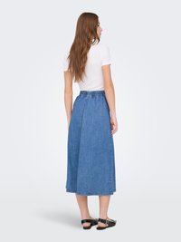 Femme aux longs cheveux bruns portant un haut blanc à manches courtes, une jupe midi en jean bleue et des chaussures plates noires, vue de profil avec le dos principalement visible.