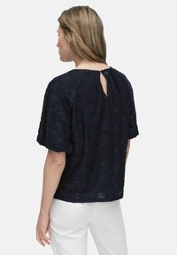 Mørkeblå bluse med korte puffede ærmer, tekstureret blomsterprint, rund halsudskæring og nøglehulslukning bagpå. Sat sammen med hvide bukser.