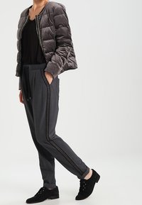 Veste matelassée grise avec fermeture éclair, haut noir, pantalon gris avec bande latérale, et chaussures noires à lacets, debout devant un fond neutre.