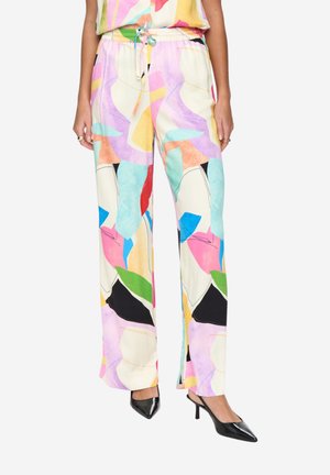 Frau trägt farbenfrohe, abstrakt bedruckte, lockere Hosen mit Kordelzugbund und schwarzen, spitz zulaufenden Absätzen, steht vor einem weißen Hintergrund.