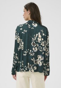 Dunkelgrün blouse mit langen Ärmeln, die eine Mischung aus cremefarbenen und blauen Blumen zeigt, mit einem lockeren Schnitt und Rückenpasse.