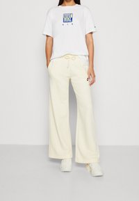 Vitt t-shirt med kort ärm och blå "Nike Air"-logotyp, kombinerat med ljusgula vida sweatpants och vita sneakers.