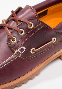 Chaussure en cuir bourgogne à bout rond, œillets dorés, lacets marron, coutures jaunes contrastées et semelle en caoutchouc beige. Présente un logo embossé sur le côté.