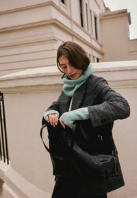 Sac fourre-tout en cuir noir à texture lisse, porté par une personne vêtue d'un manteau gris et d'une écharpe tricotée bleu clair, en train de récupérer des objets.