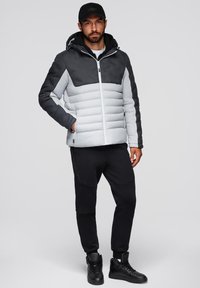 Puffjacka med grå och svart färgsättning, med dragkedjestängning, justerbar huva och ribbade ärmslut. Modellen bär svarta byxor och sneakers.