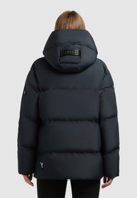 Puffer jacket in dark navy, featuring a large hood, stitched panels, and a branded patch on the back. Smooth, water-resistant fabric.