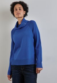 Frau trägt einen leuchtend blauen Strickpullover mit Knopfleiste am Hals und dunklen Jeans und steht vor einem schlichten, neutralen Hintergrund.