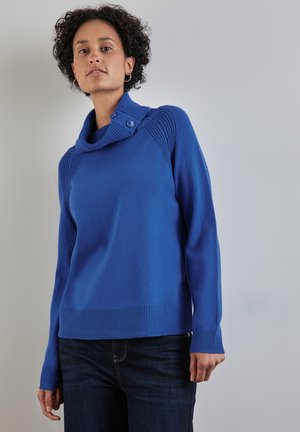 Vrouw in een felblauwe gebreide trui met knopen in de kraag en donkere jeans, staand tegen een effen neutrale achtergrond.