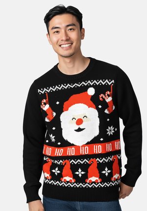 Junger Mann lächelt und trägt einen schwarzen Weihnachtspullover mit einem Weihnachtsmann-Gesicht, Handschuhen, Zuckerstangen, Schneeflocken und dem Text "HO HO HO".
