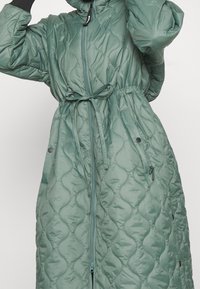 Donna che indossa un lungo cappotto verde trapuntato con cintura in vita, zip frontale e tasche con automatici, mentre aggiusta il cappuccio con una mano guantata.