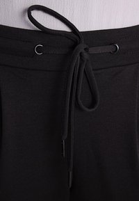 Čierny elastický pás so šnúrkou, ktorý má hladký povrch a strieborné kovové očká na nastavenie, s plochým čiernym šnúrkou.