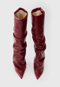 Botas altas, holgadas y de punta afilada en cuero rojo profundo, de pie sobre un fondo blanco.