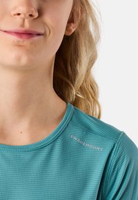 Nærbillede af en persons nedre ansigt og hals iført en teal tekstureret skjorte med "Swedemount" trykt nær skulderen.