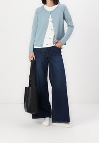 Helles blaues Cardigan mit Knopfverschluss, getragen über einem weißen Hemd mit kleinen Mustern. Dunkelblaue weite Jeans und eine schwarze Tragetasche. Weiße Sneakers.